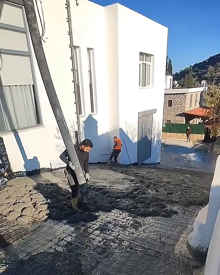 Beyaz villanın önünde beton pompası hortumuyla zemin dökümü yapan işçiler ve arka planda ağaçlı yamaç