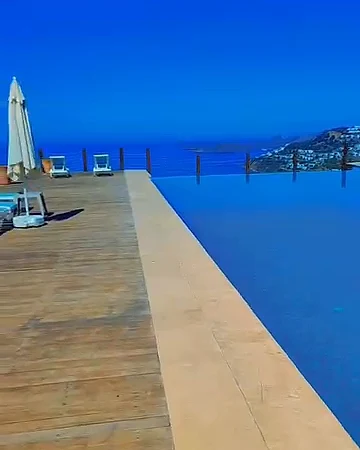 Ahşap teraslı sonsuzluk havuzu, kenarda şemsiye ve şezlonglar, uzakta deniz ve kıyı yerleşimi