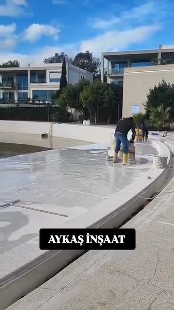 Boş havuzun açık gri zemininde sarı çizmeli usta, arkada iki katlı konutlar ve ağaçlar