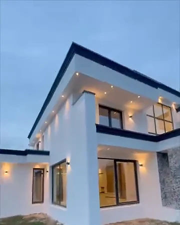Beyaz, iki katlı, düz çatılı modern villa; alacakaranlıkta yanan duvar aydınlatmaları ve geniş cam kapılar